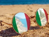 Okulary przeciwsłoneczne na plaży z odbiciem włoskich krajobrazów w szkłach, ilustrujące letnie kursy języka włoskiego w szkole Porto Felice.