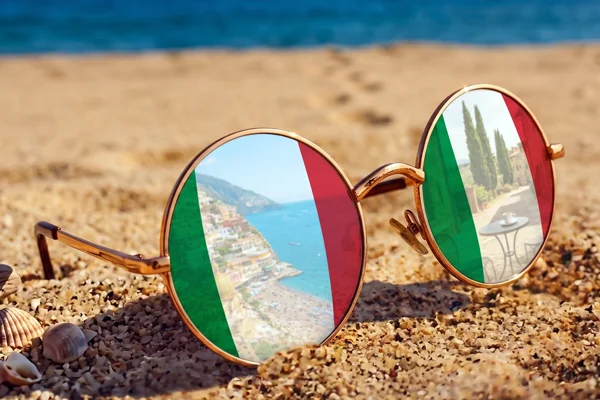 Okulary przeciwsłoneczne na plaży z odbiciem włoskich krajobrazów w szkłach, ilustrujące letnie kursy języka włoskiego w szkole Porto Felice.